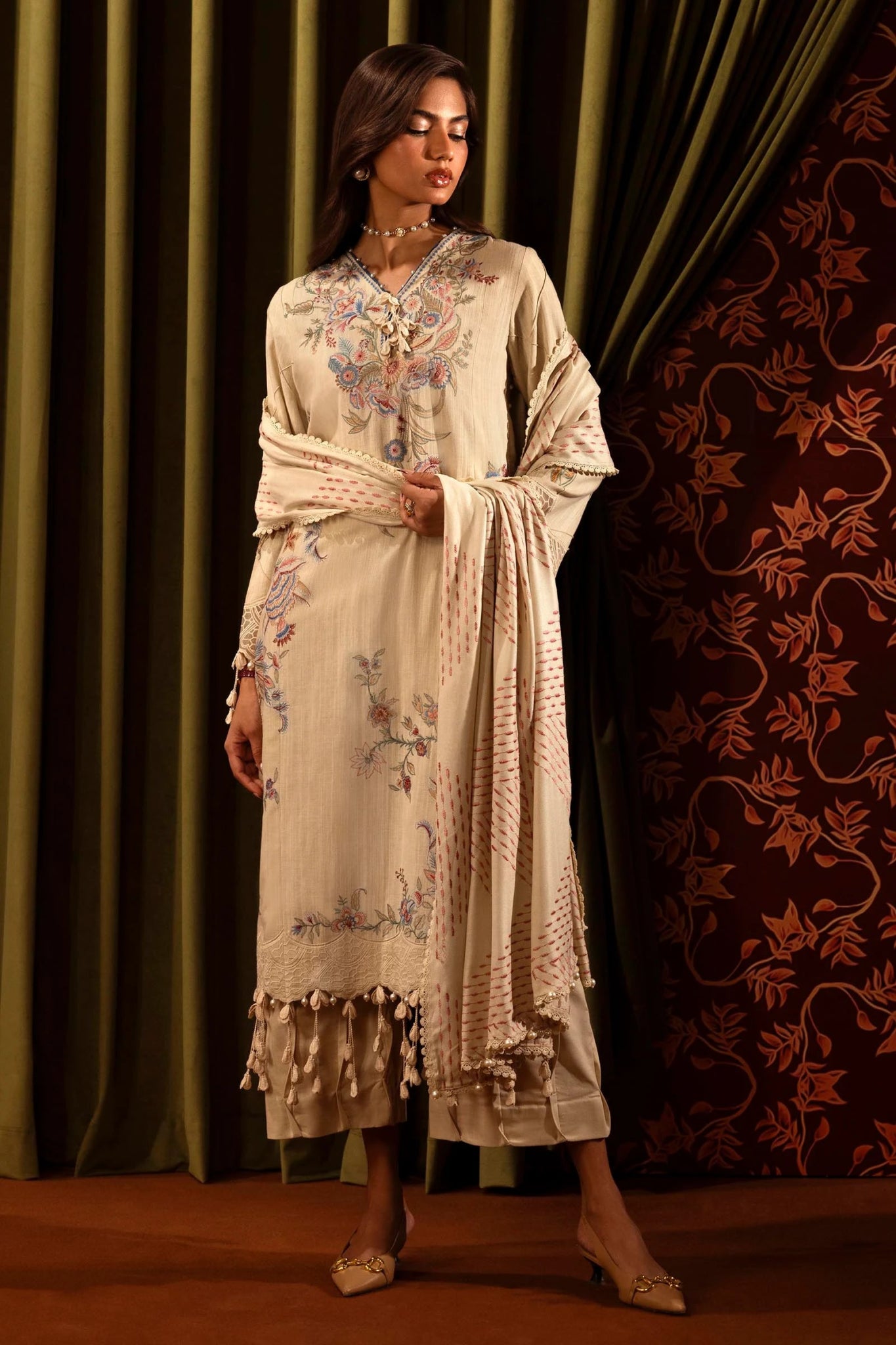 A model showcases a three - piece outfit from the Sana Safinaz Muzlin Winter '24 Collection. The design includes an embroidered front kali on slub in three pieces, paired with a dyed slub back and matching embroidered sleeves. The ensemble is completed with a delicate embroidered organza kuful, a detailed embroidered shawl, and coordinating dyed cotton pants. Ideal for a luxurious and sophisticated look.