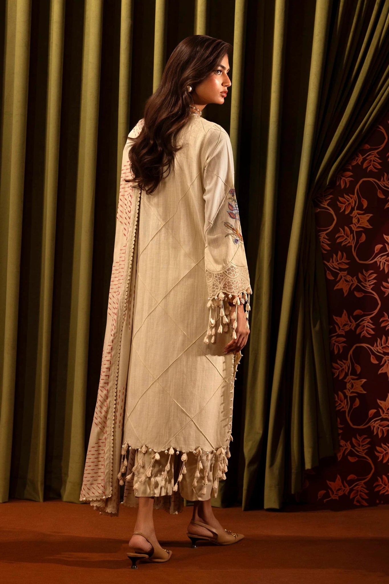 A model showcases a three - piece outfit from the Sana Safinaz Muzlin Winter '24 Collection. The design includes an embroidered front kali on slub in three pieces, paired with a dyed slub back and matching embroidered sleeves. The ensemble is completed with a delicate embroidered organza kuful, a detailed embroidered shawl, and coordinating dyed cotton pants. Ideal for a luxurious and sophisticated look.