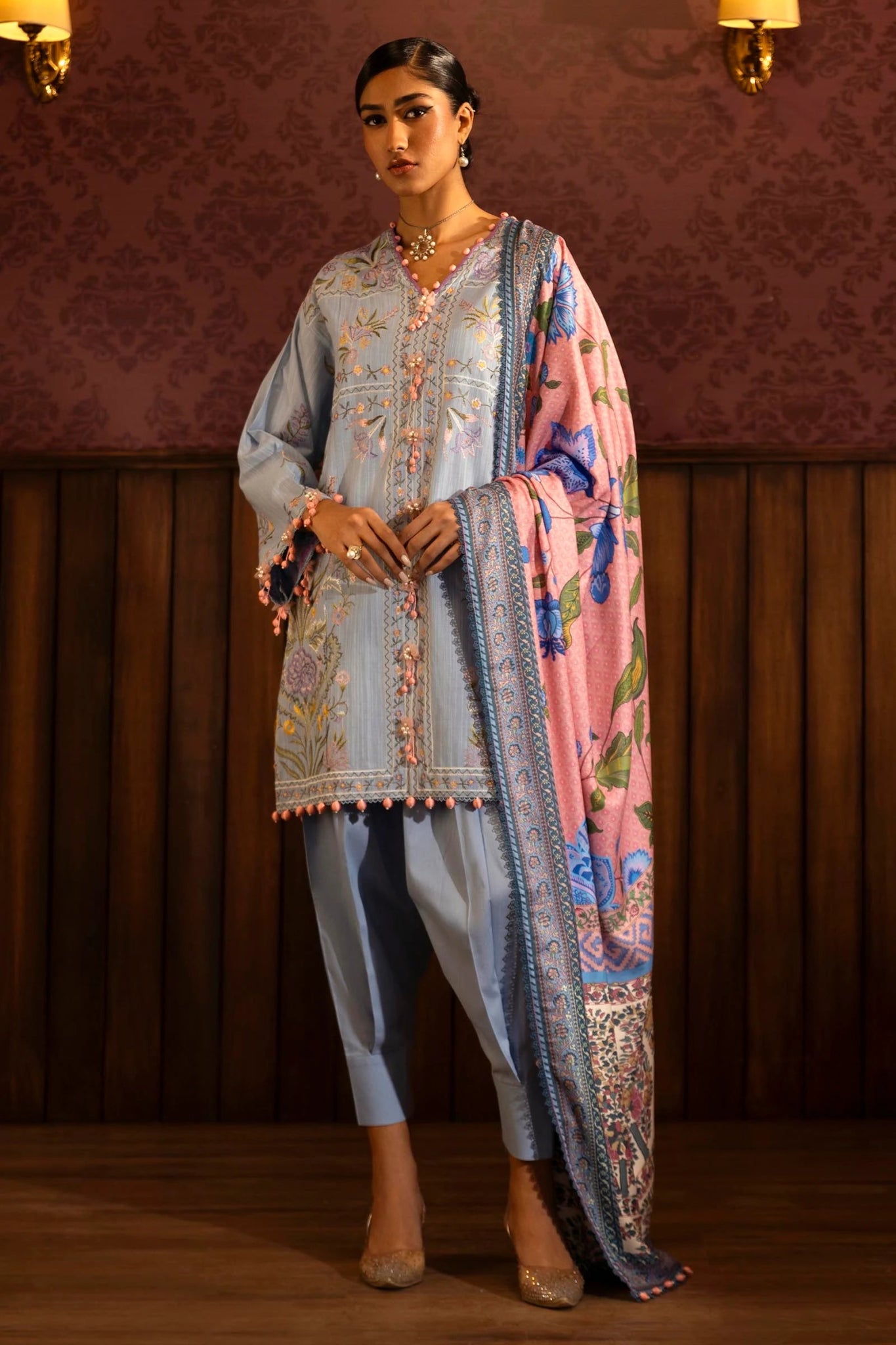 Model wearing a 3 - piece pastel blue embroidered suit featuring a slub front kali, embroidered sleeves, and a digitally printed shawl with floral patterns. Paired with dyed cotton pants for a chic look. Part of the Sana Safinaz Muzlin Winter 2024 collection, available online in the UK