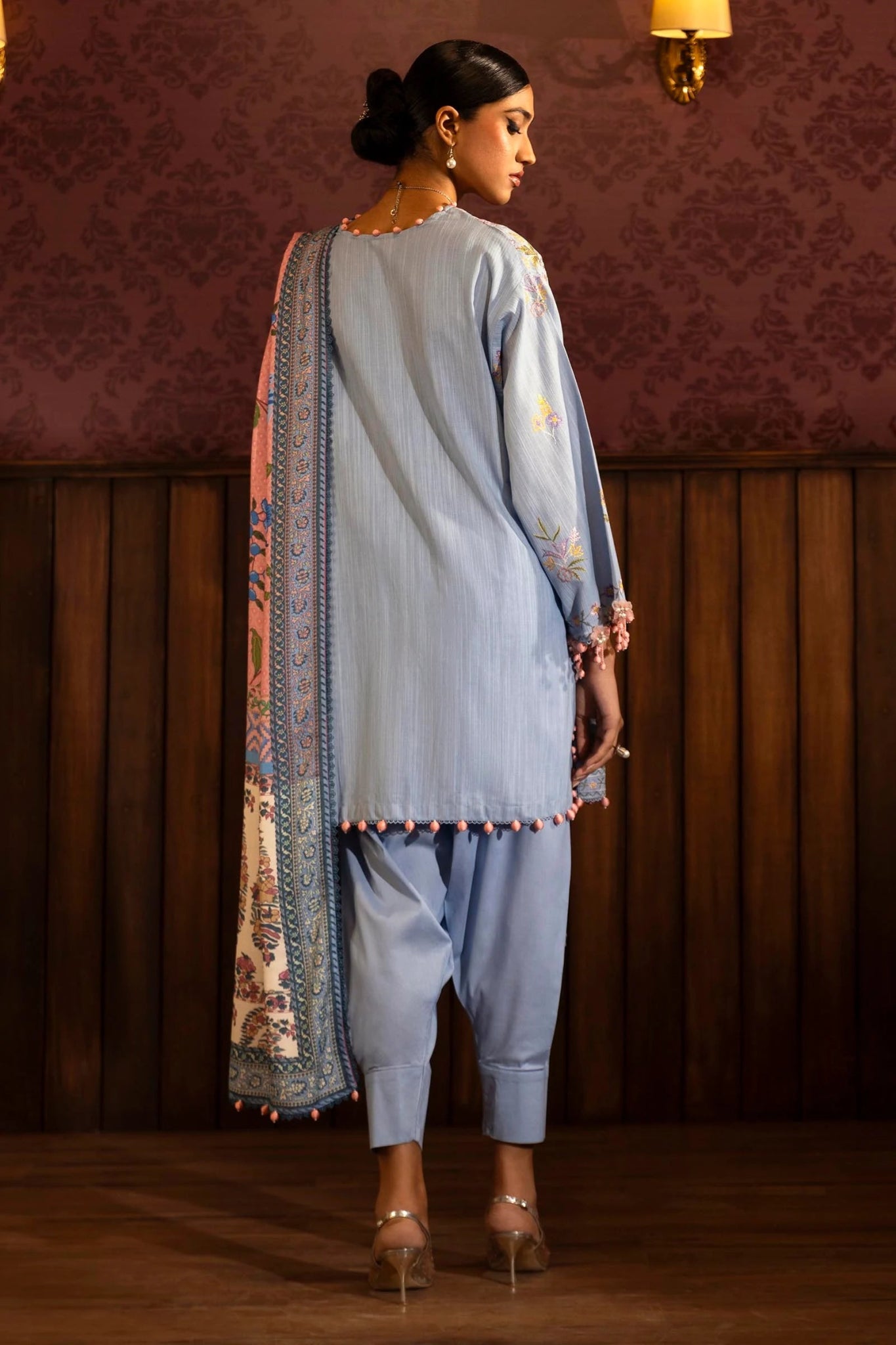 Model wearing a 3 - piece pastel blue embroidered suit featuring a slub front kali, embroidered sleeves, and a digitally printed shawl with floral patterns. Paired with dyed cotton pants for a chic look. Part of the Sana Safinaz Muzlin Winter 2024 collection, available online in the UK