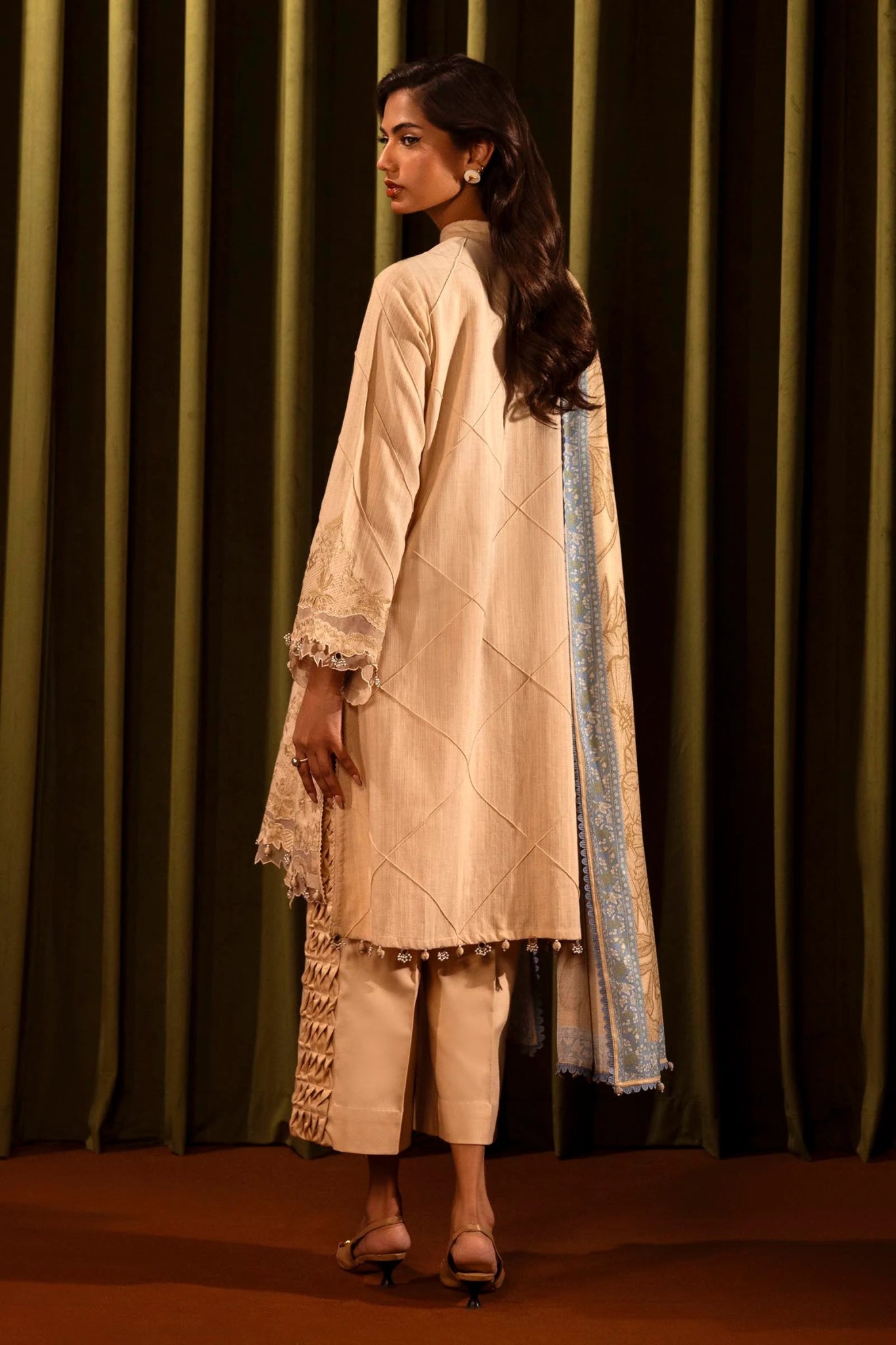 Model wearing a 3 - piece beige embroidered suit featuring a slub front kali, embroidered sleeves with organza details, and a printed shawl with blue borders. Paired with dyed cotton pants for a chic look. Part of the Sana Safinaz Muzlin Winter 2024 collection, available online in the UK.