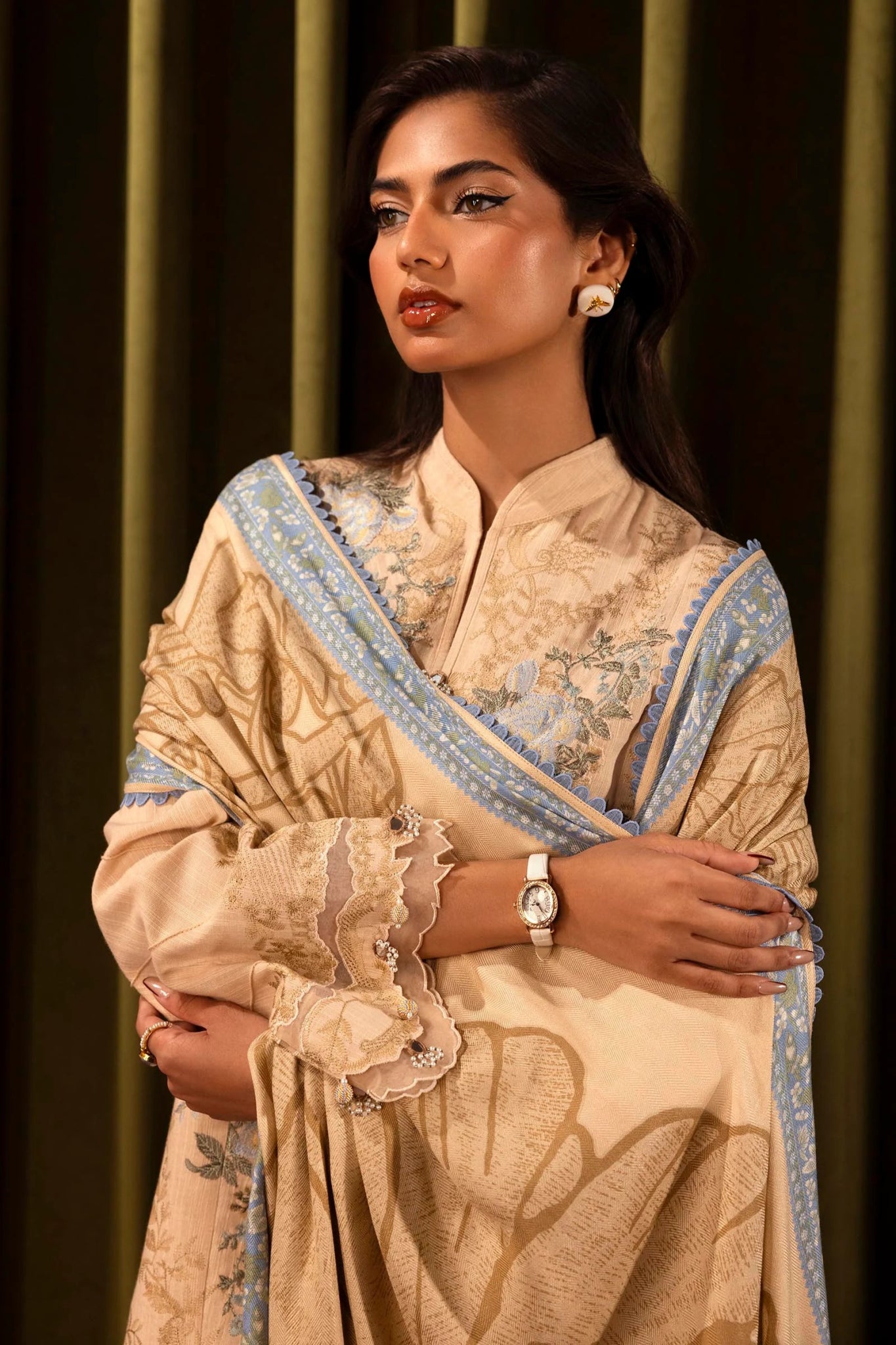 Model wearing a 3 - piece beige embroidered suit featuring a slub front kali, embroidered sleeves with organza details, and a printed shawl with blue borders. Paired with dyed cotton pants for a chic look. Part of the Sana Safinaz Muzlin Winter 2024 collection, available online in the UK.