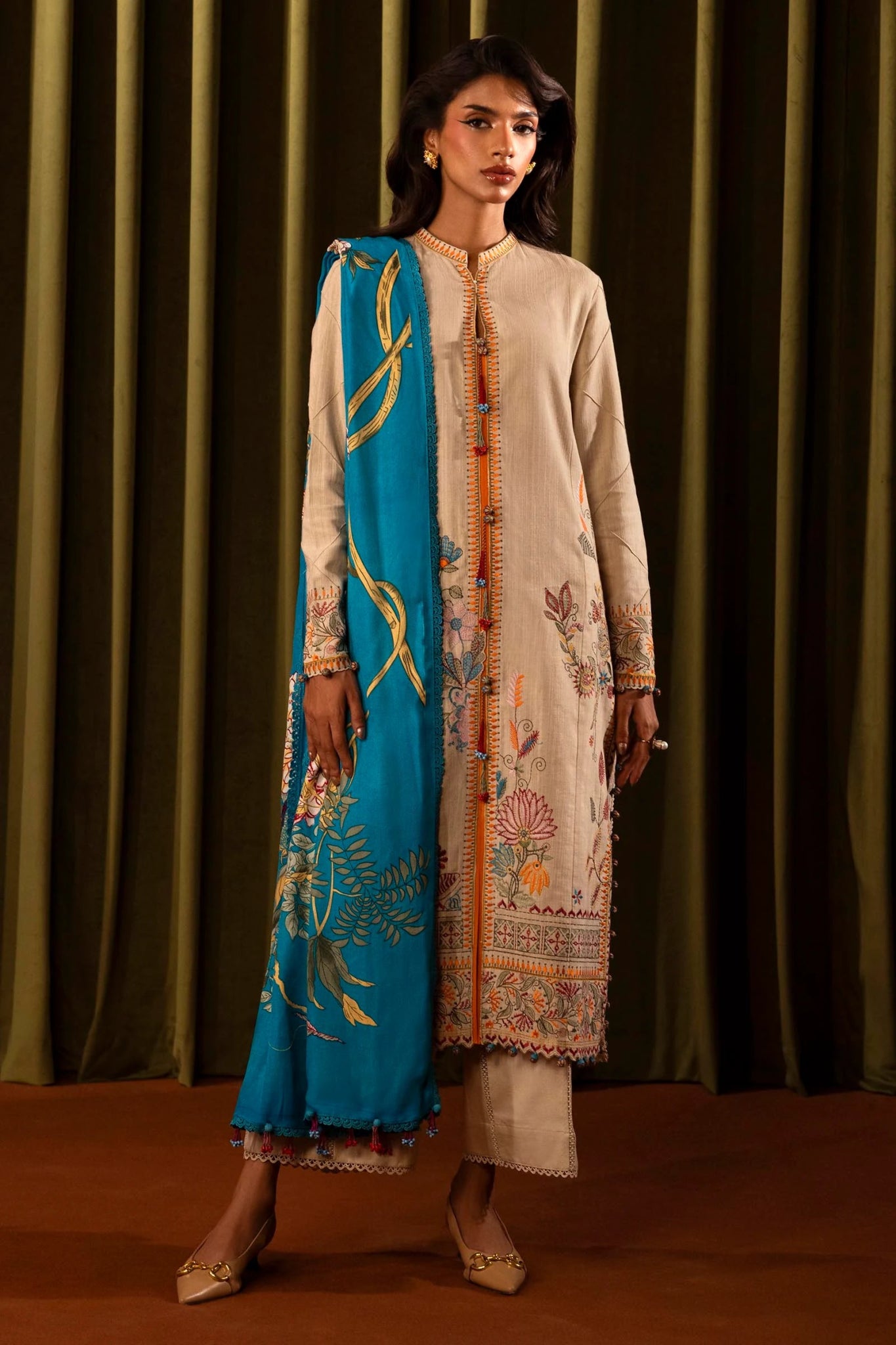 Model wearing a 3 - piece beige embroidered suit featuring a slub front kali, embroidered sleeves, and a vibrant blue printed shawl. Paired with dyed cotton pants for a sophisticated look. Part of the Sana Safinaz Muzlin Winter 2024 collection, available online in the UK