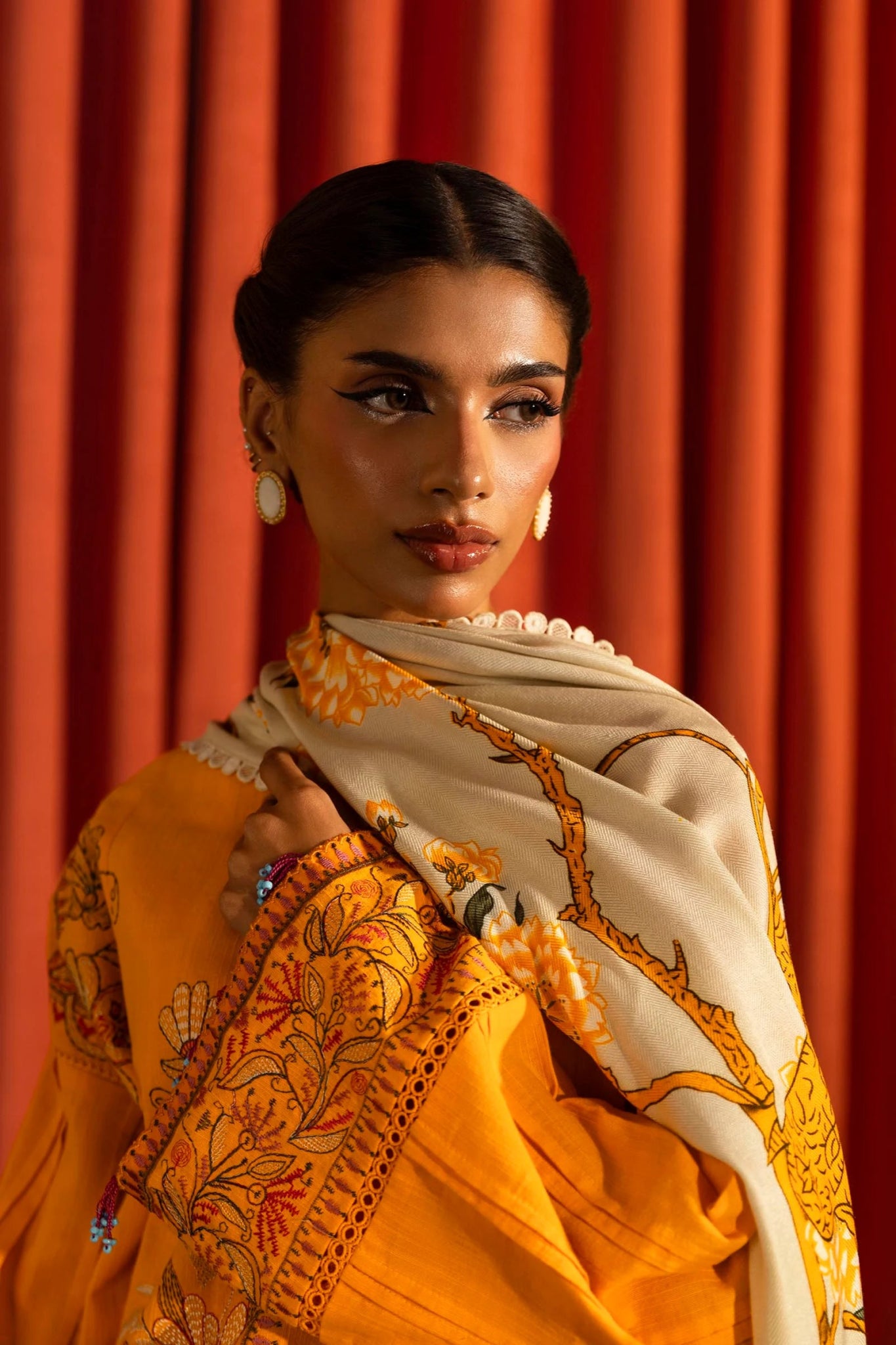 Model wearing a 3 - piece bright orange embroidered suit featuring a slub front kali, embroidered sleeves, and a printed shawl with floral accents. Paired with dyed cotton pants for a vibrant look. Part of the Sana Safinaz Muzlin Winter 2024 collection, available online in the UK.