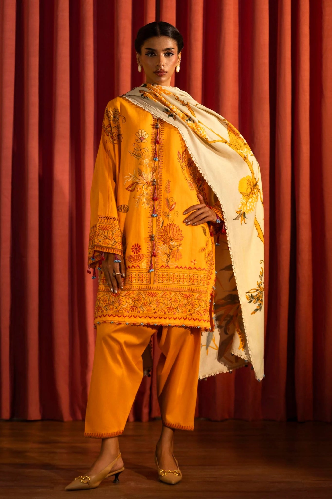 Model wearing a 3 - piece bright orange embroidered suit featuring a slub front kali, embroidered sleeves, and a printed shawl with floral accents. Paired with dyed cotton pants for a vibrant look. Part of the Sana Safinaz Muzlin Winter 2024 collection, available online in the UK.