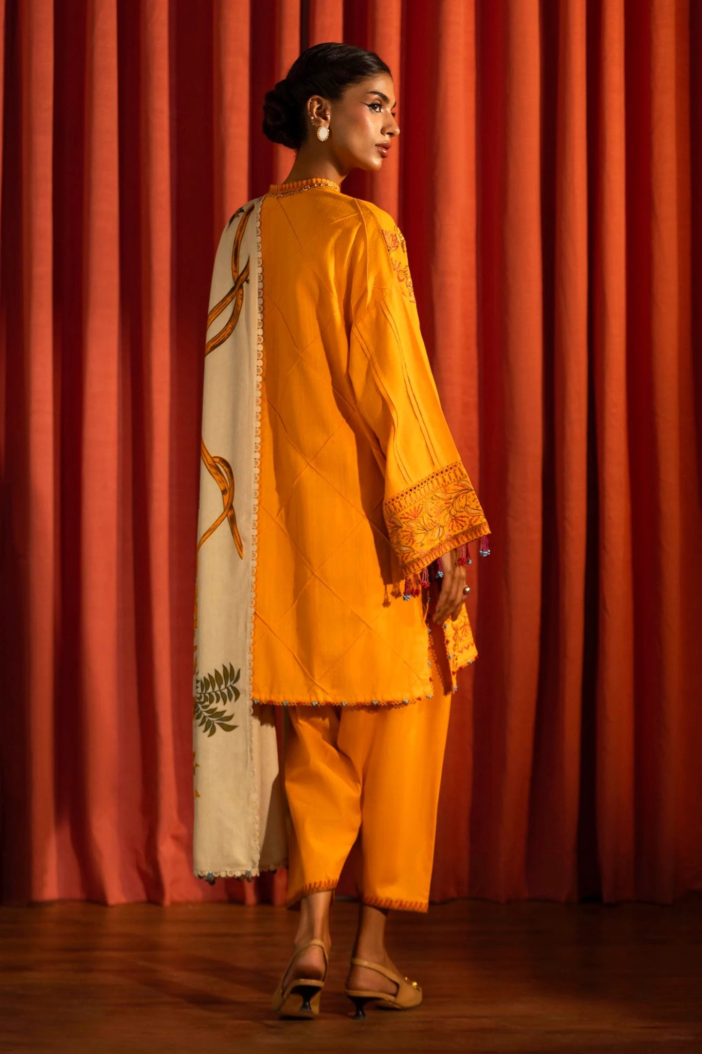 Model wearing a 3 - piece bright orange embroidered suit featuring a slub front kali, embroidered sleeves, and a printed shawl with floral accents. Paired with dyed cotton pants for a vibrant look. Part of the Sana Safinaz Muzlin Winter 2024 collection, available online in the UK.