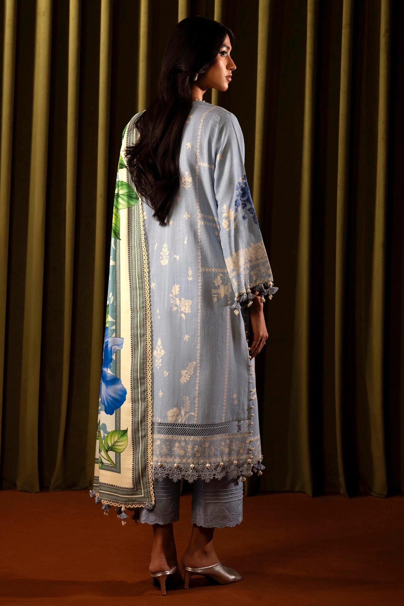 Model wearing a 3 - piece light blue embroidered suit featuring a slub front kali, printed back and sleeves, and a digital printed shawl with floral patterns. Paired with dyed cotton pants for an elegant look. Part of the Sana Safinaz Muzlin Winter 2024 collection, available online in the UK.