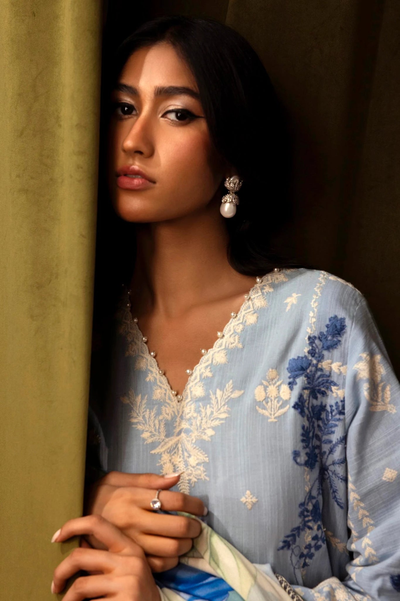 Model wearing a 3 - piece light blue embroidered suit featuring a slub front kali, printed back and sleeves, and a digital printed shawl with floral patterns. Paired with dyed cotton pants for an elegant look. Part of the Sana Safinaz Muzlin Winter 2024 collection, available online in the UK.