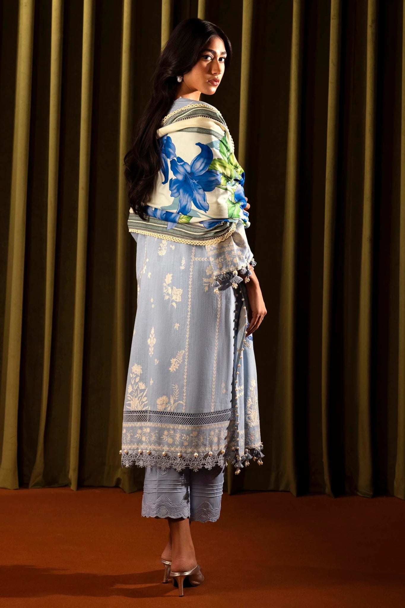 Model wearing a 3 - piece light blue embroidered suit featuring a slub front kali, printed back and sleeves, and a digital printed shawl with floral patterns. Paired with dyed cotton pants for an elegant look. Part of the Sana Safinaz Muzlin Winter 2024 collection, available online in the UK.