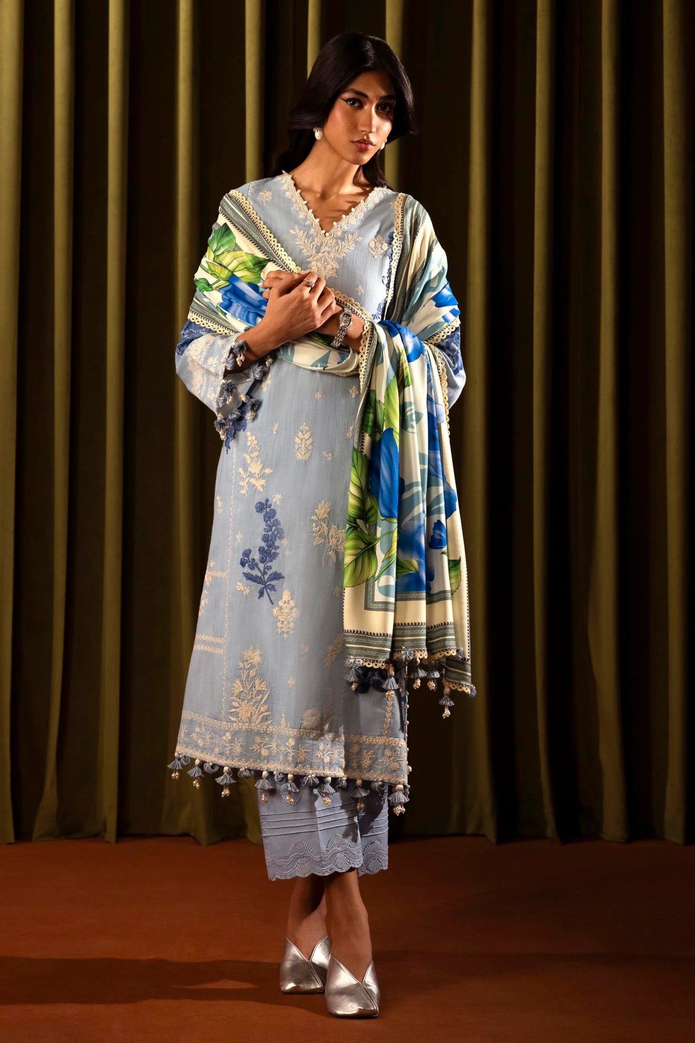 Model wearing a 3 - piece light blue embroidered suit featuring a slub front kali, printed back and sleeves, and a digital printed shawl with floral patterns. Paired with dyed cotton pants for an elegant look. Part of the Sana Safinaz Muzlin Winter 2024 collection, available online in the UK.