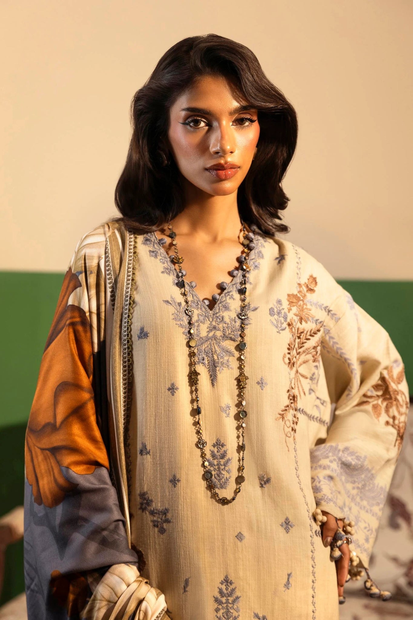 Model wearing a 3 - piece beige embroidered suit featuring a slub front kali, printed back and sleeves, and a digital printed shawl with bold floral patterns. Paired with dyed cotton pants for an elegant look. Part of the Sana Safinaz Muzlin Winter 2024 collection, available online in the UK.