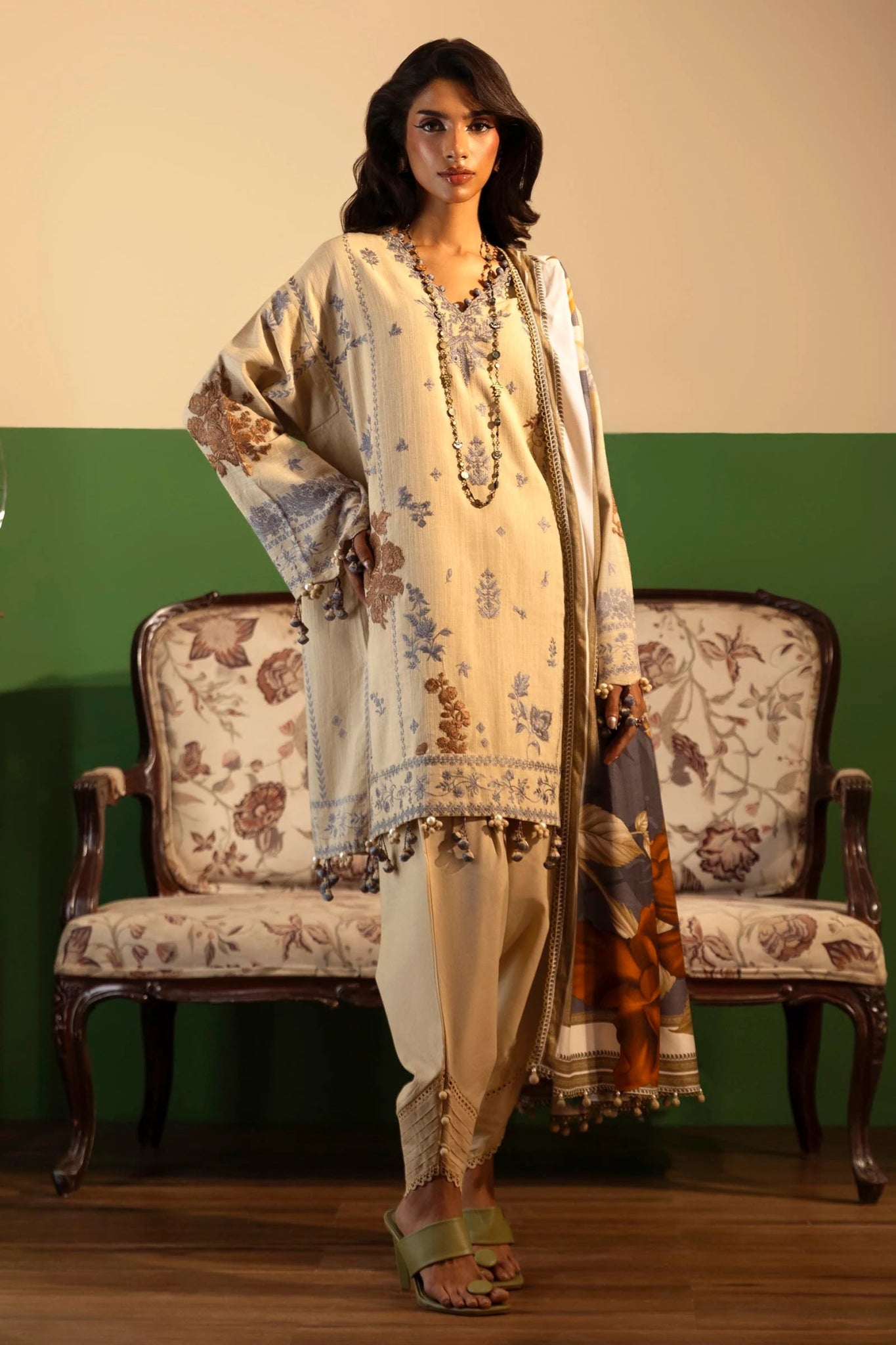 Model wearing a 3 - piece beige embroidered suit featuring a slub front kali, printed back and sleeves, and a digital printed shawl with bold floral patterns. Paired with dyed cotton pants for an elegant look. Part of the Sana Safinaz Muzlin Winter 2024 collection, available online in the UK.