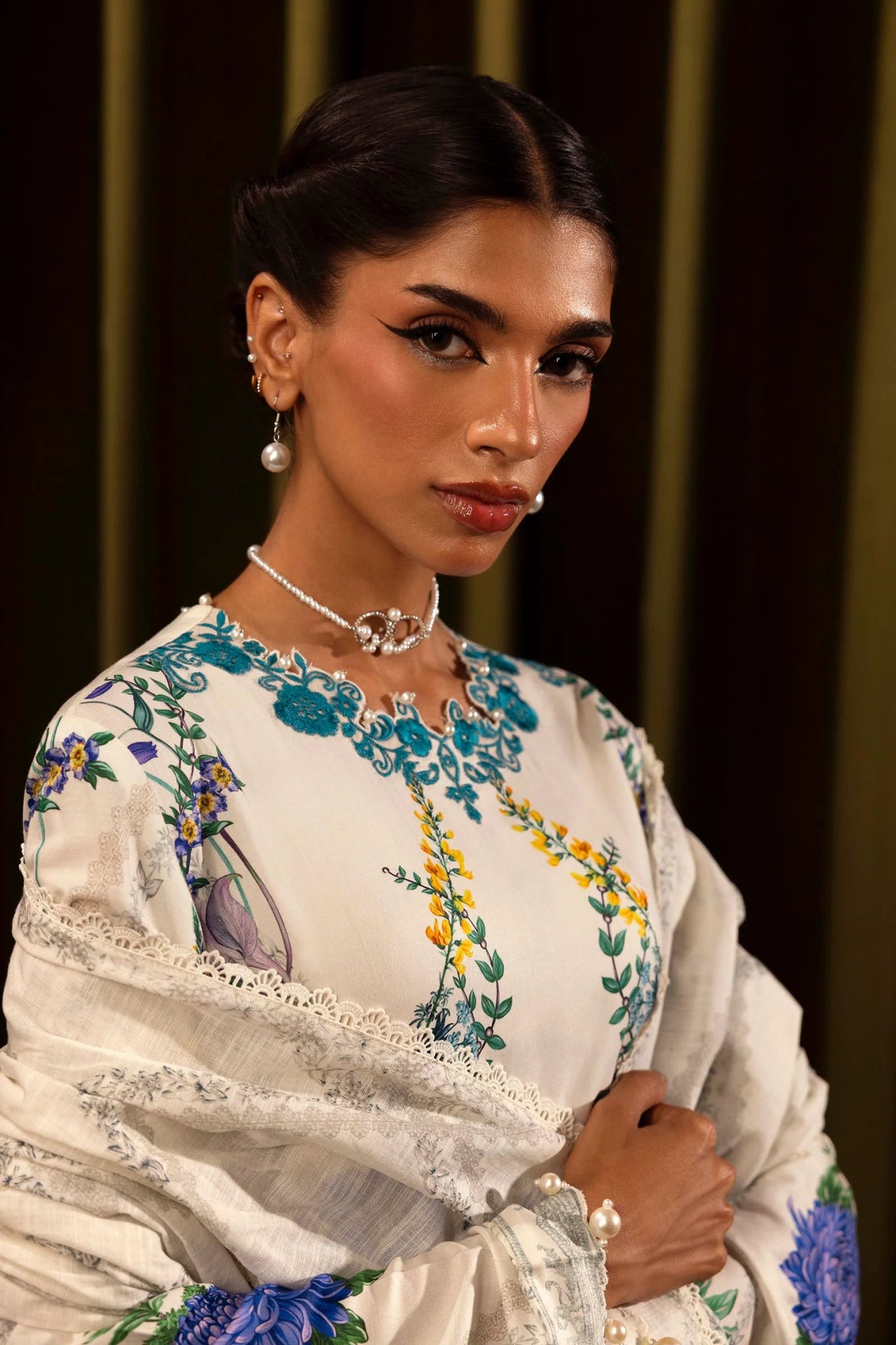 Model wearing a 3 - piece white suit featuring a digitally printed linen front, back, and sleeves with blue floral patterns. Styled with an embroidered organza neckline and border, a printed khaddar dupatta, and dyed cotton pants for a chic look. Part of the Sana Safinaz Muzlin Winter 2024 collection, available online in the UK.