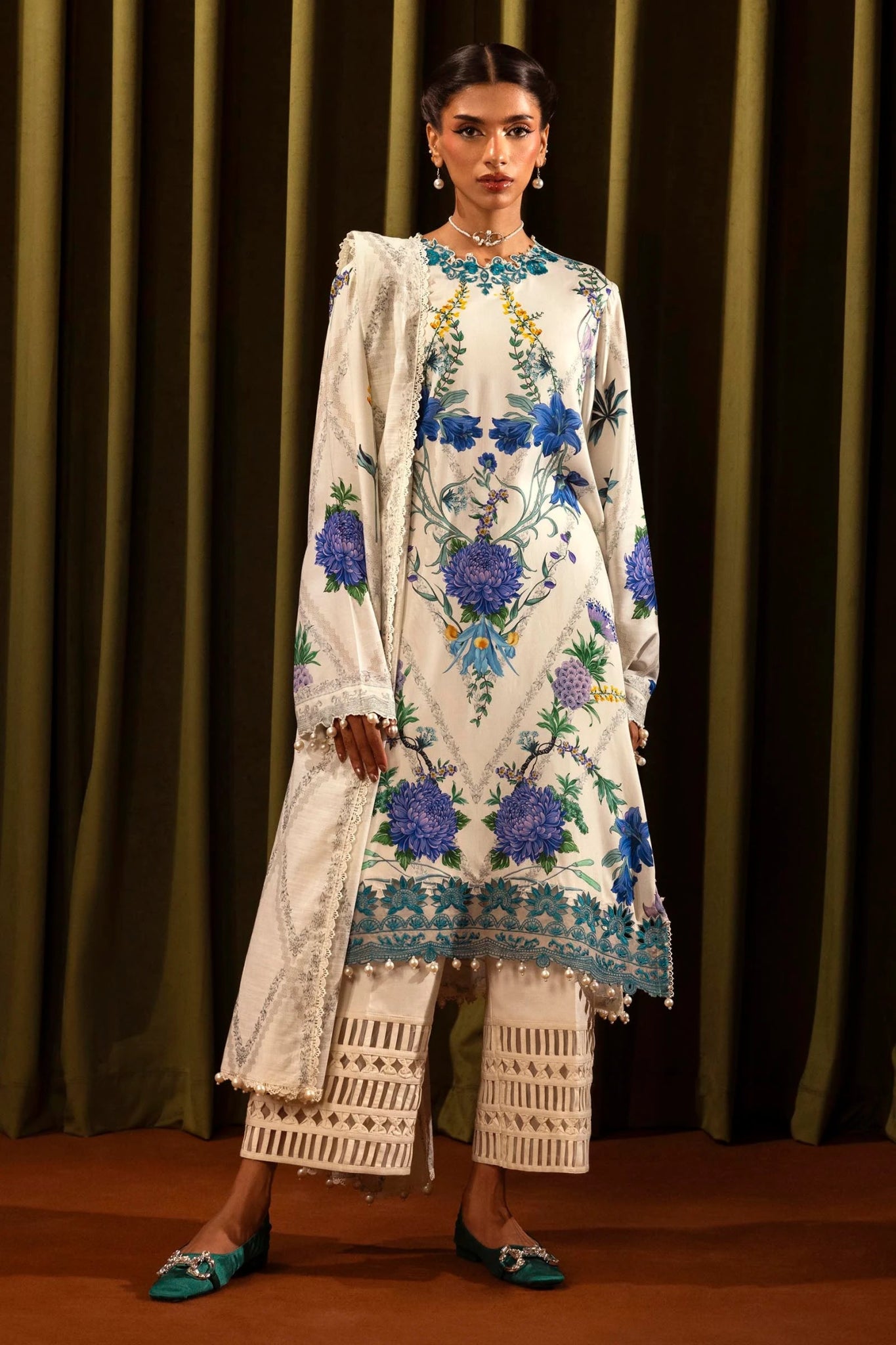 Model wearing a 3 - piece white suit featuring a digitally printed linen front, back, and sleeves with blue floral patterns. Styled with an embroidered organza neckline and border, a printed khaddar dupatta, and dyed cotton pants for a chic look. Part of the Sana Safinaz Muzlin Winter 2024 collection, available online in the UK.