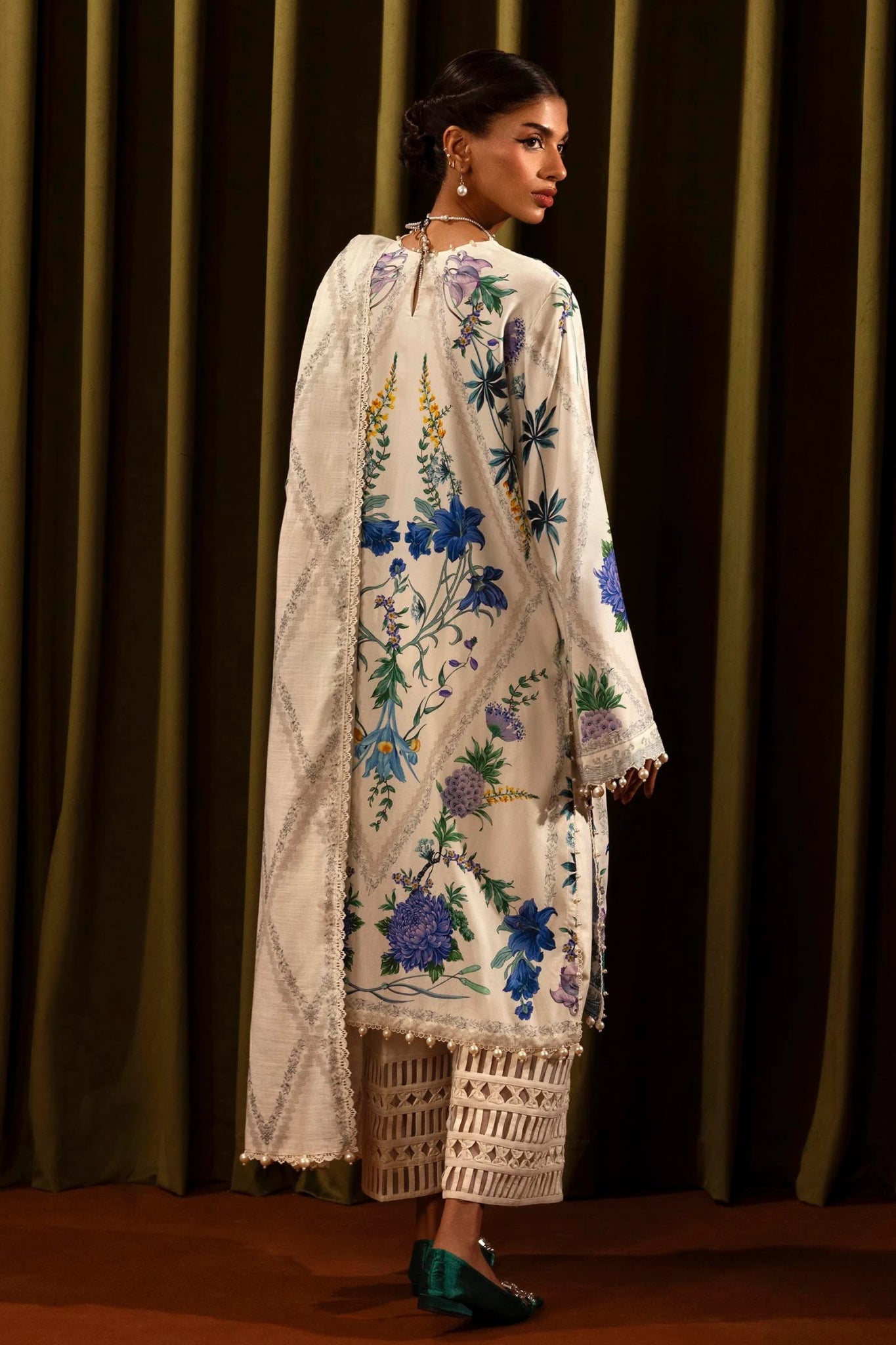 Model wearing a 3 - piece white suit featuring a digitally printed linen front, back, and sleeves with blue floral patterns. Styled with an embroidered organza neckline and border, a printed khaddar dupatta, and dyed cotton pants for a chic look. Part of the Sana Safinaz Muzlin Winter 2024 collection, available online in the UK.