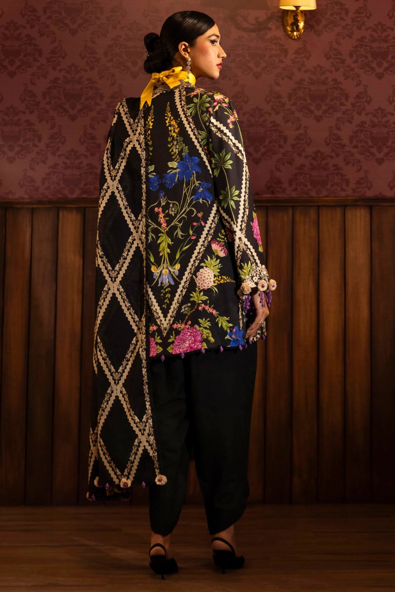 Model wearing a 3 - piece black suit featuring a digitally printed linen front, back, and sleeves with vibrant floral patterns. Styled with an embroidered organza neckline and border, a printed khaddar dupatta, and dyed cotton pants for an elegant look. Part of the Sana Safinaz Muzlin Winter 2024 collection, available online in the UK.