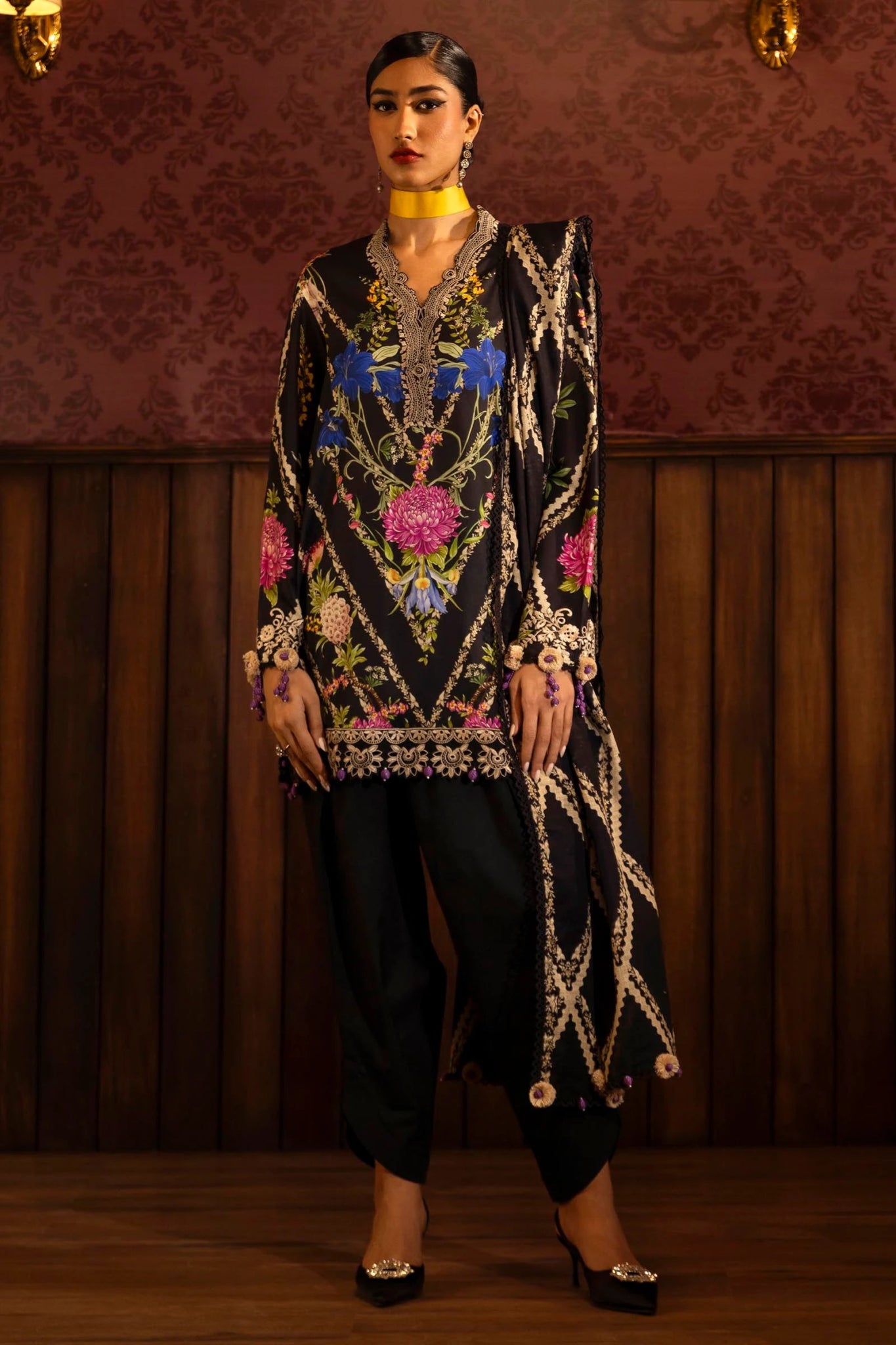 Model wearing a 3 - piece black suit featuring a digitally printed linen front, back, and sleeves with vibrant floral patterns. Styled with an embroidered organza neckline and border, a printed khaddar dupatta, and dyed cotton pants for an elegant look. Part of the Sana Safinaz Muzlin Winter 2024 collection, available online in the UK.