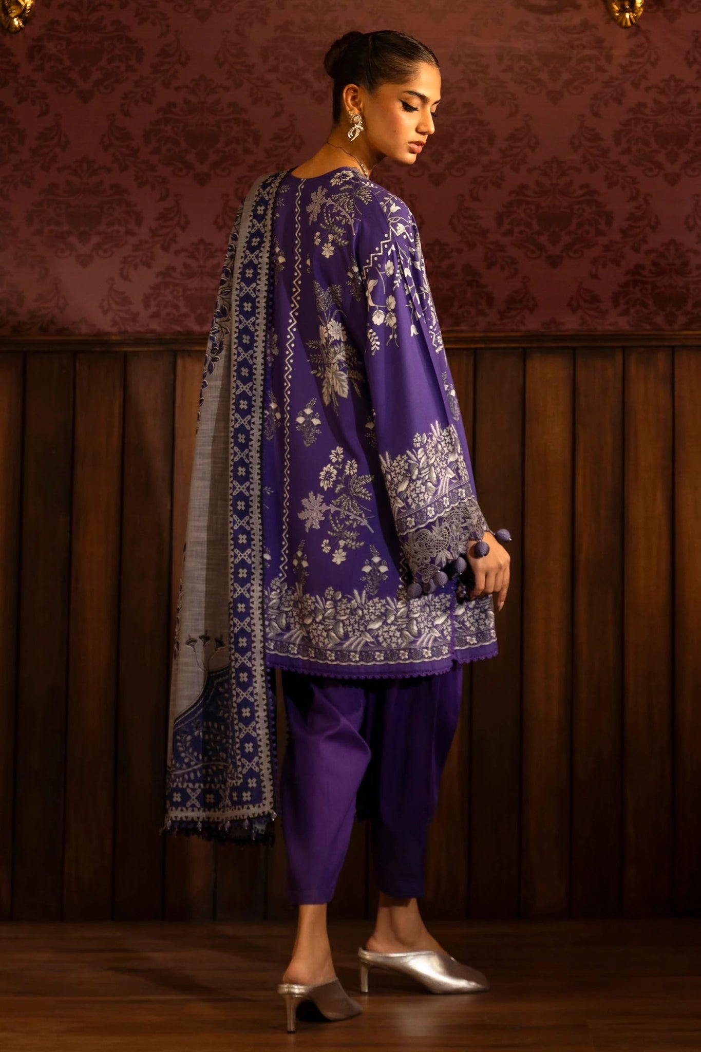 Model wearing a 3 - piece deep purple suit featuring a digitally printed linen front, back, and sleeves with intricate patterns. Styled with an embroidered organza neckline and border, a printed khaddar dupatta, and dyed cotton pants for an elegant look. Part of the Sana Safinaz Muzlin Winter 2024 collection, available online in the UK.