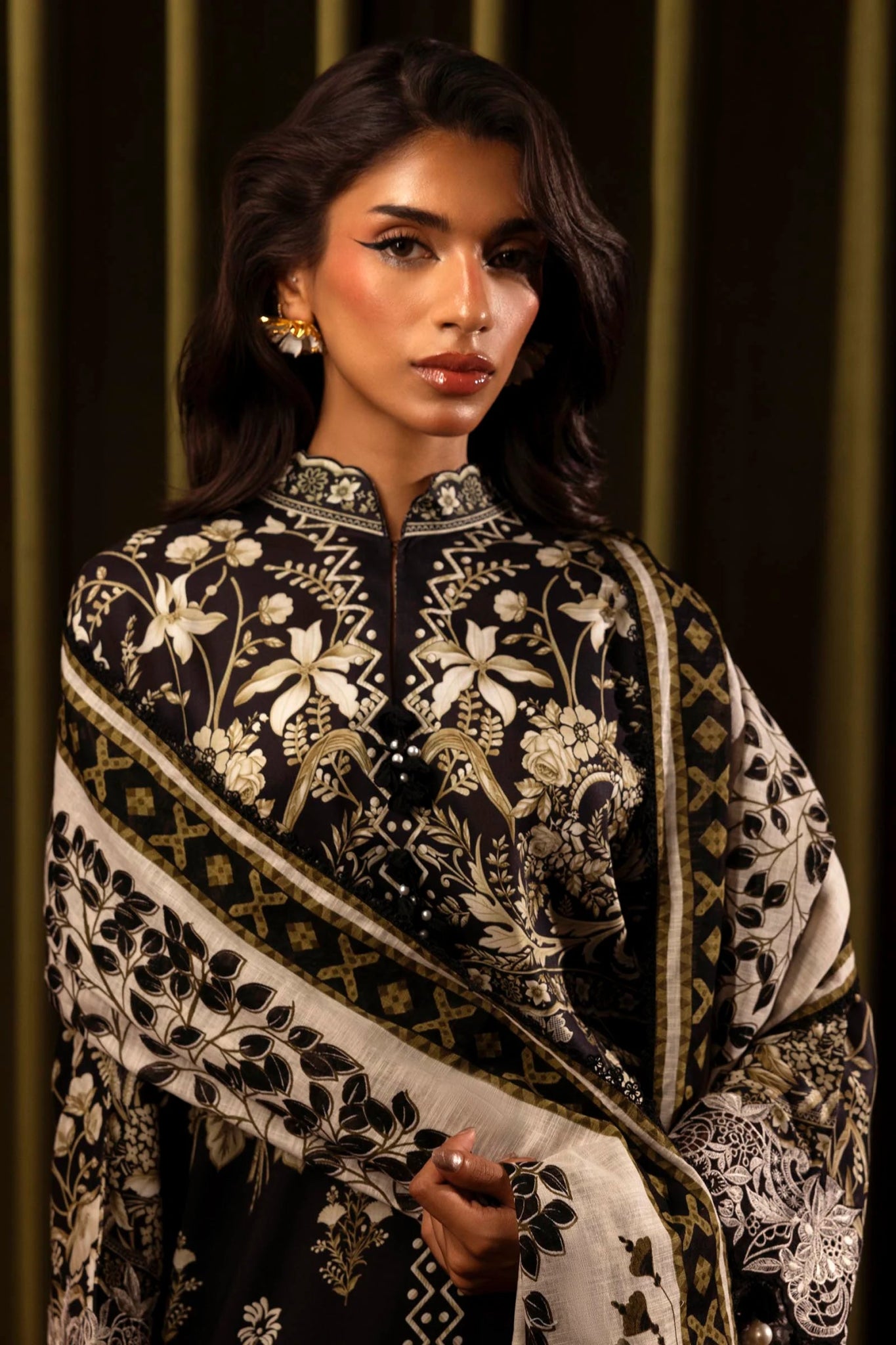 Model wearing a 3 - piece black suit featuring a digitally printed linen front, back, and sleeves with intricate beige floral patterns. Styled with an embroidered organza neckline and border, a printed khaddar dupatta, and dyed cotton pants for an elegant look. Part of the Sana Safinaz Muzlin Winter 2024 collection, available online in the UK.