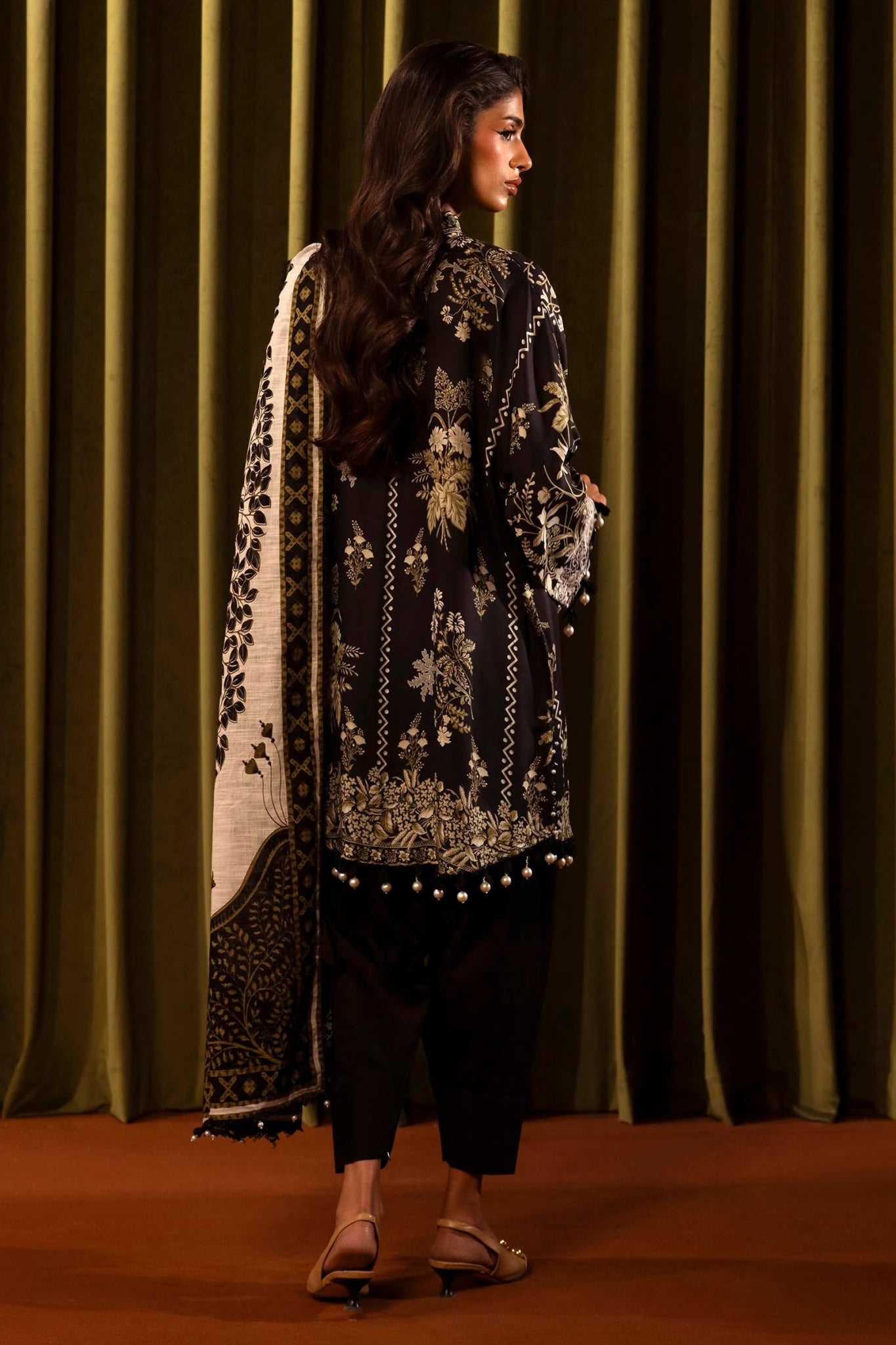 Model wearing a 3 - piece black suit featuring a digitally printed linen front, back, and sleeves with intricate beige floral patterns. Styled with an embroidered organza neckline and border, a printed khaddar dupatta, and dyed cotton pants for an elegant look. Part of the Sana Safinaz Muzlin Winter 2024 collection, available online in the UK.