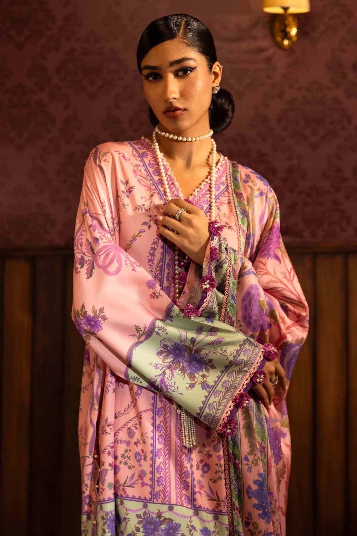 Model wearing a 3 - piece pink suit featuring a digitally printed linen front, back, and sleeves with intricate purple and green floral patterns. Styled with an embroidered organza border, a printed linen dupatta, and dyed cotton pants with embroidered accents. Part of the Sana Safinaz Muzlin Winter 2024 collection, available online in the UK.