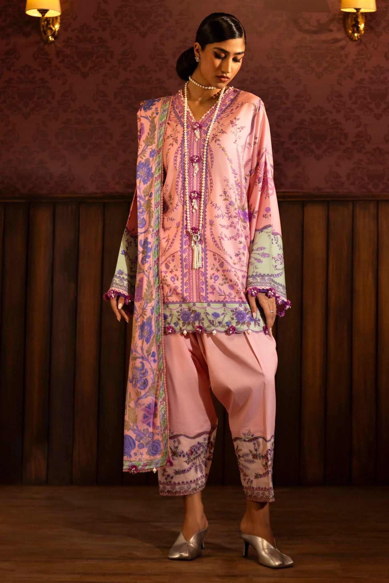 Model wearing a 3 - piece pink suit featuring a digitally printed linen front, back, and sleeves with intricate purple and green floral patterns. Styled with an embroidered organza border, a printed linen dupatta, and dyed cotton pants with embroidered accents. Part of the Sana Safinaz Muzlin Winter 2024 collection, available online in the UK.