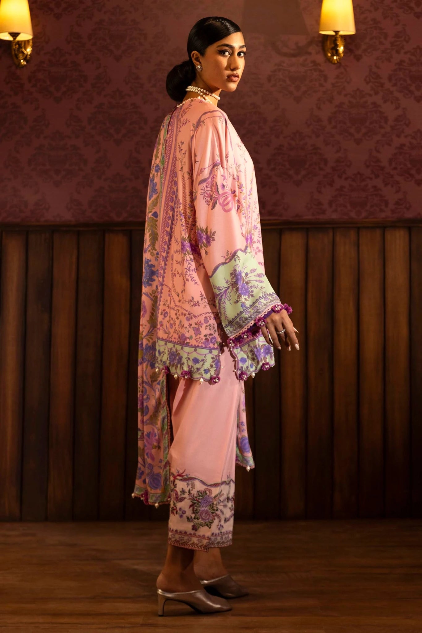 Model wearing a 3 - piece pink suit featuring a digitally printed linen front, back, and sleeves with intricate purple and green floral patterns. Styled with an embroidered organza border, a printed linen dupatta, and dyed cotton pants with embroidered accents. Part of the Sana Safinaz Muzlin Winter 2024 collection, available online in the UK.