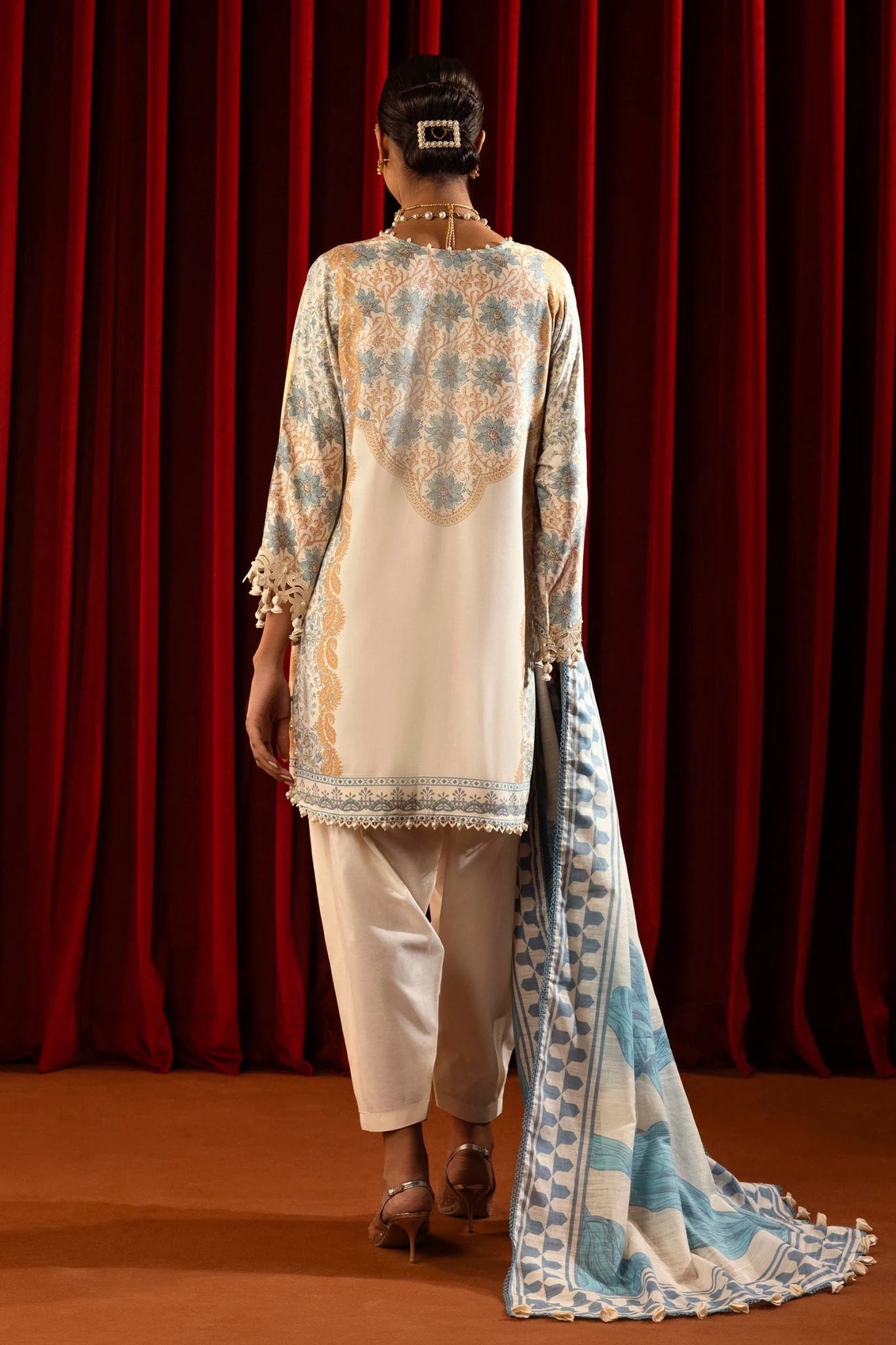 Model wearing a 3 - piece cream suit featuring a digitally printed linen front, back, and sleeves with blue and golden accents. Styled with an embroidered organza neckline and sleeve borders, a printed khaddar dupatta, and dyed cotton pants for an elegant look. Part of the Sana Safinaz Muzlin Winter 2024 collection, available online in the UK.