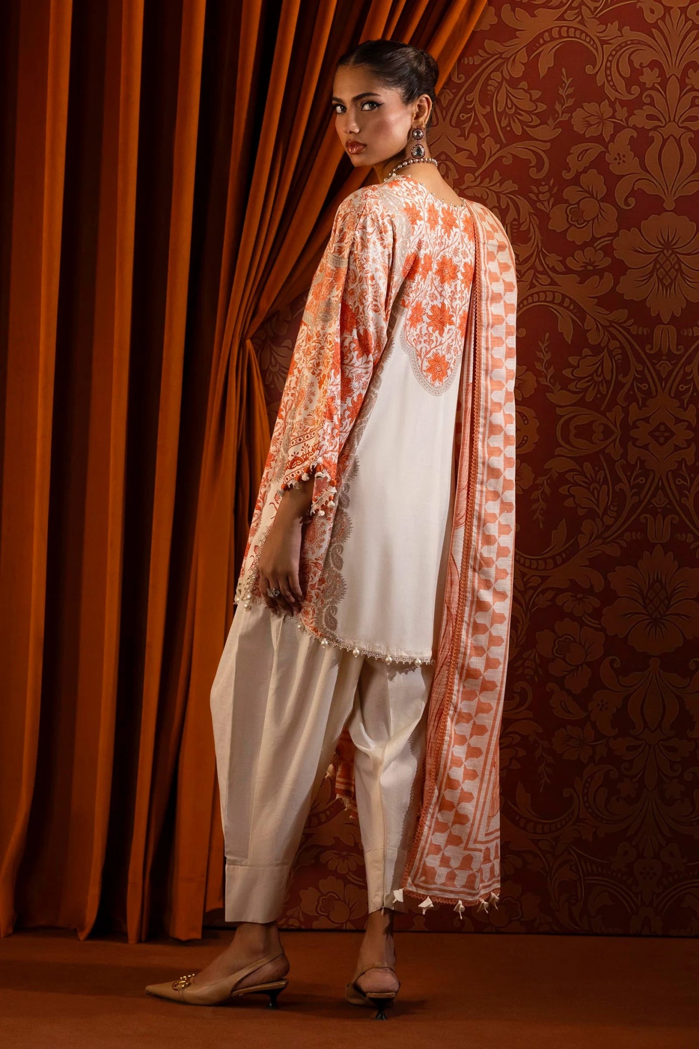 Model wearing a 3 - piece cream and orange suit featuring a digitally printed linen front, back, and sleeves with intricate orange floral patterns. Styled with an embroidered organza neckline and sleeve borders, a printed khaddar dupatta, and dyed cotton pants for an elegant look. Part of the Sana Safinaz Muzlin Winter 2024 collection, available online in the UK.