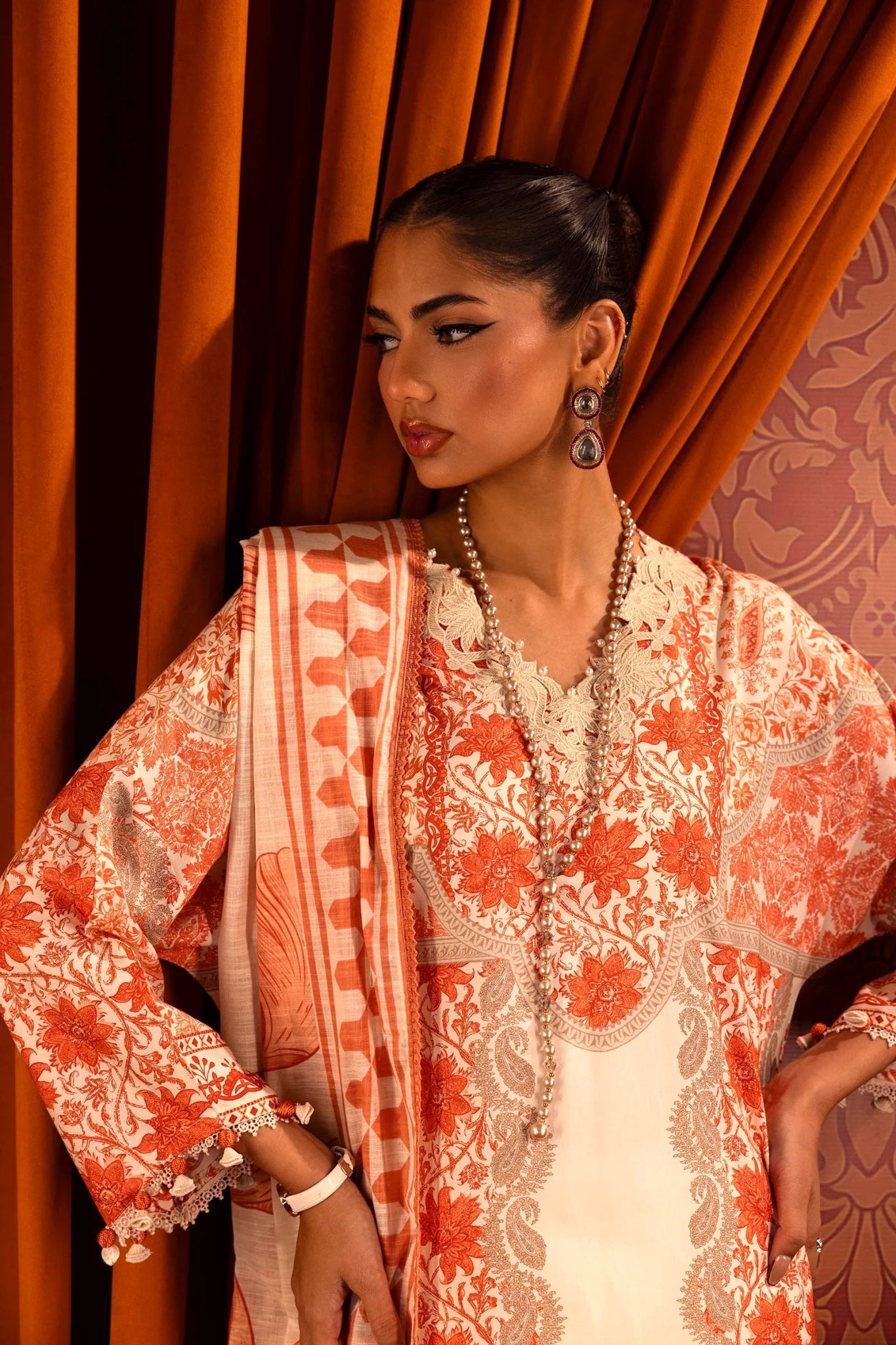 Model wearing a 3 - piece cream and orange suit featuring a digitally printed linen front, back, and sleeves with intricate orange floral patterns. Styled with an embroidered organza neckline and sleeve borders, a printed khaddar dupatta, and dyed cotton pants for an elegant look. Part of the Sana Safinaz Muzlin Winter 2024 collection, available online in the UK.