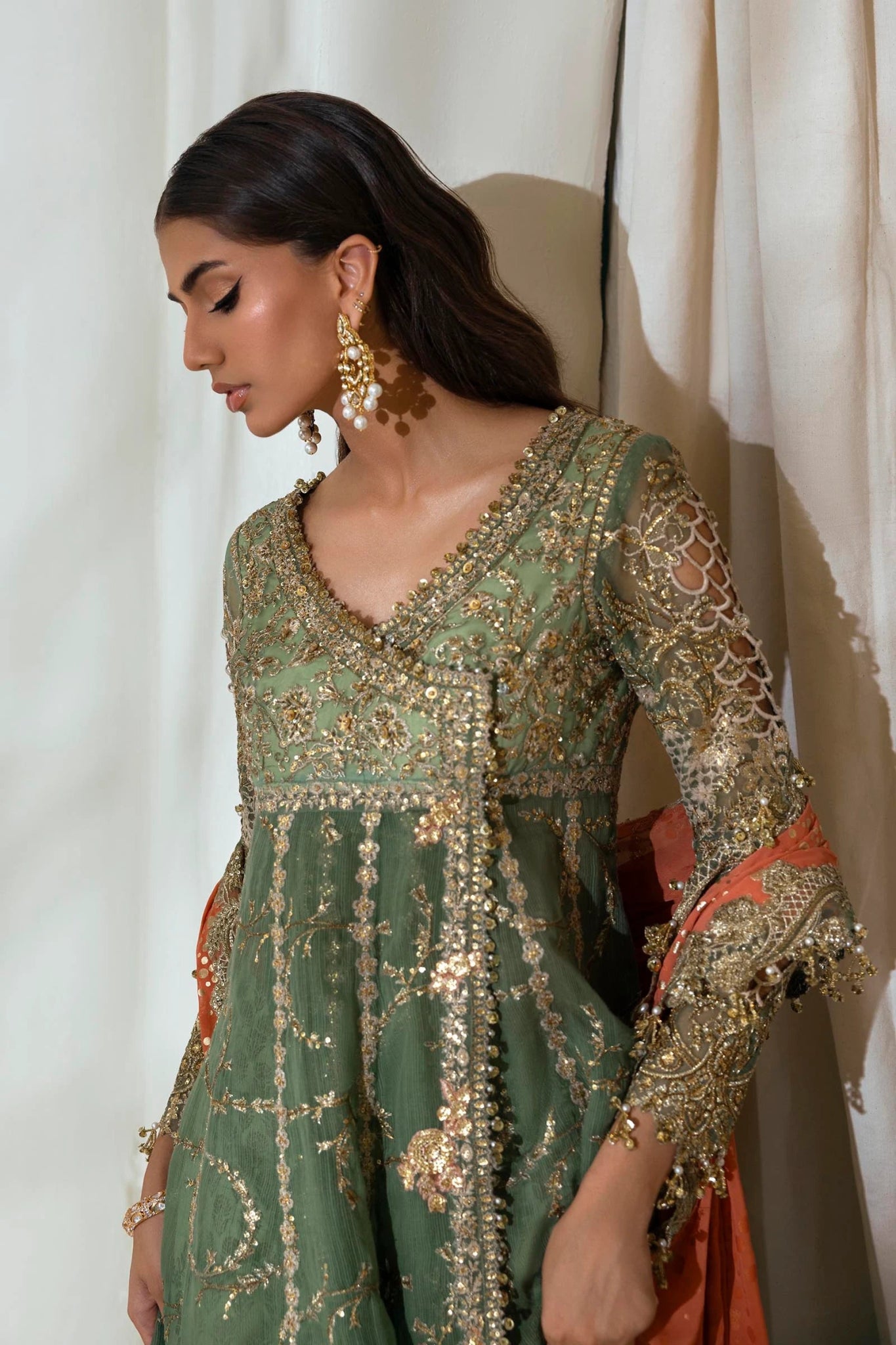 Model wearing Sana Safinaz NURA Festive'24 Vol. II N242 - 006 - 3CX dress, featuring a green ensemble with gold embellishments and an orange dupatta. Perfect for weddings and available as Pakistani clothes online in the UK.