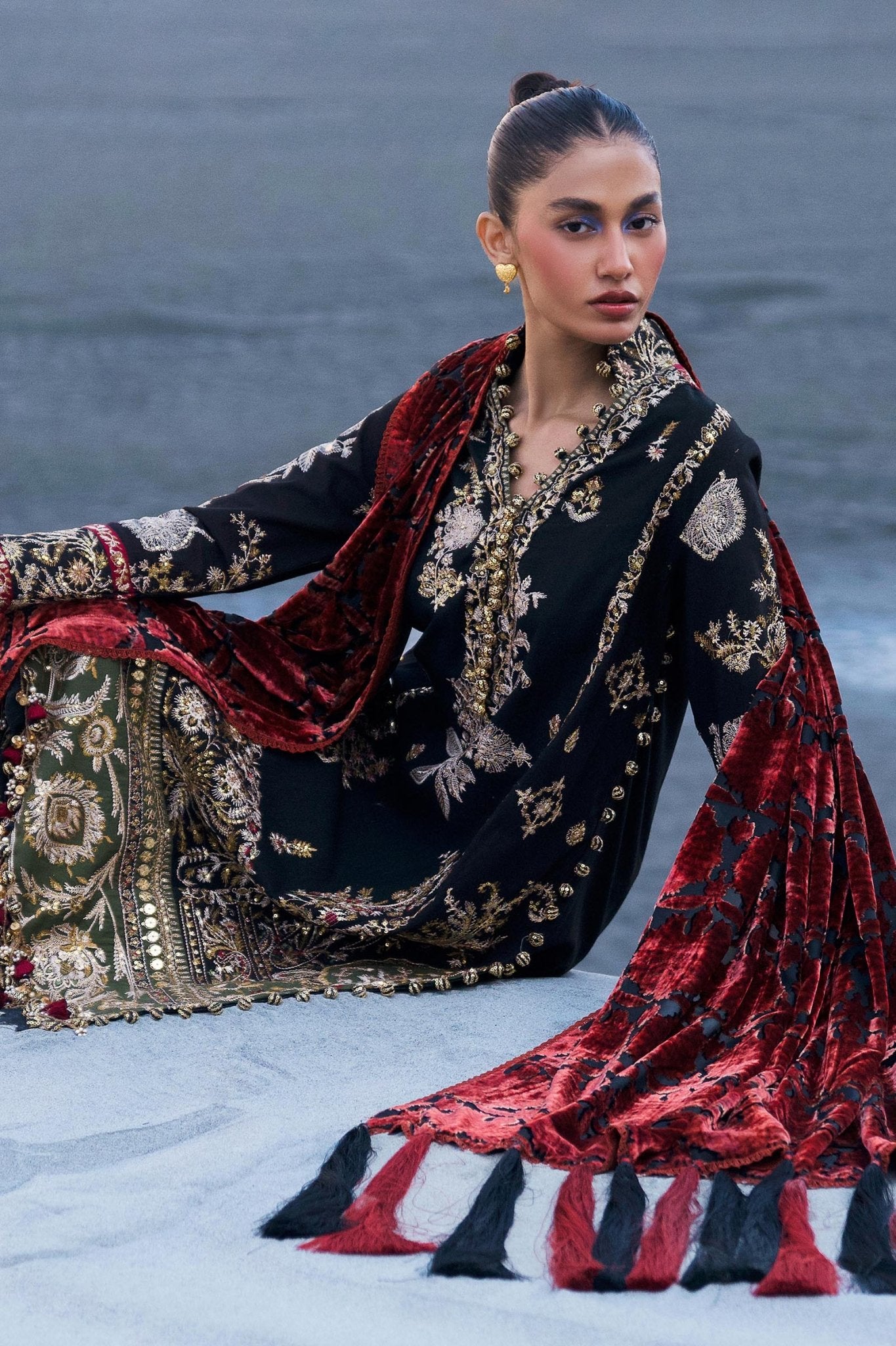 Model wearing Sana Safinaz Winter Luxury '24 S241 - 001A - 3CP, a black and gold embroidered outfit with intricate patterns and a bold red velvet shawl. Perfect for winter, available for Pakistani clothes online in the UK.