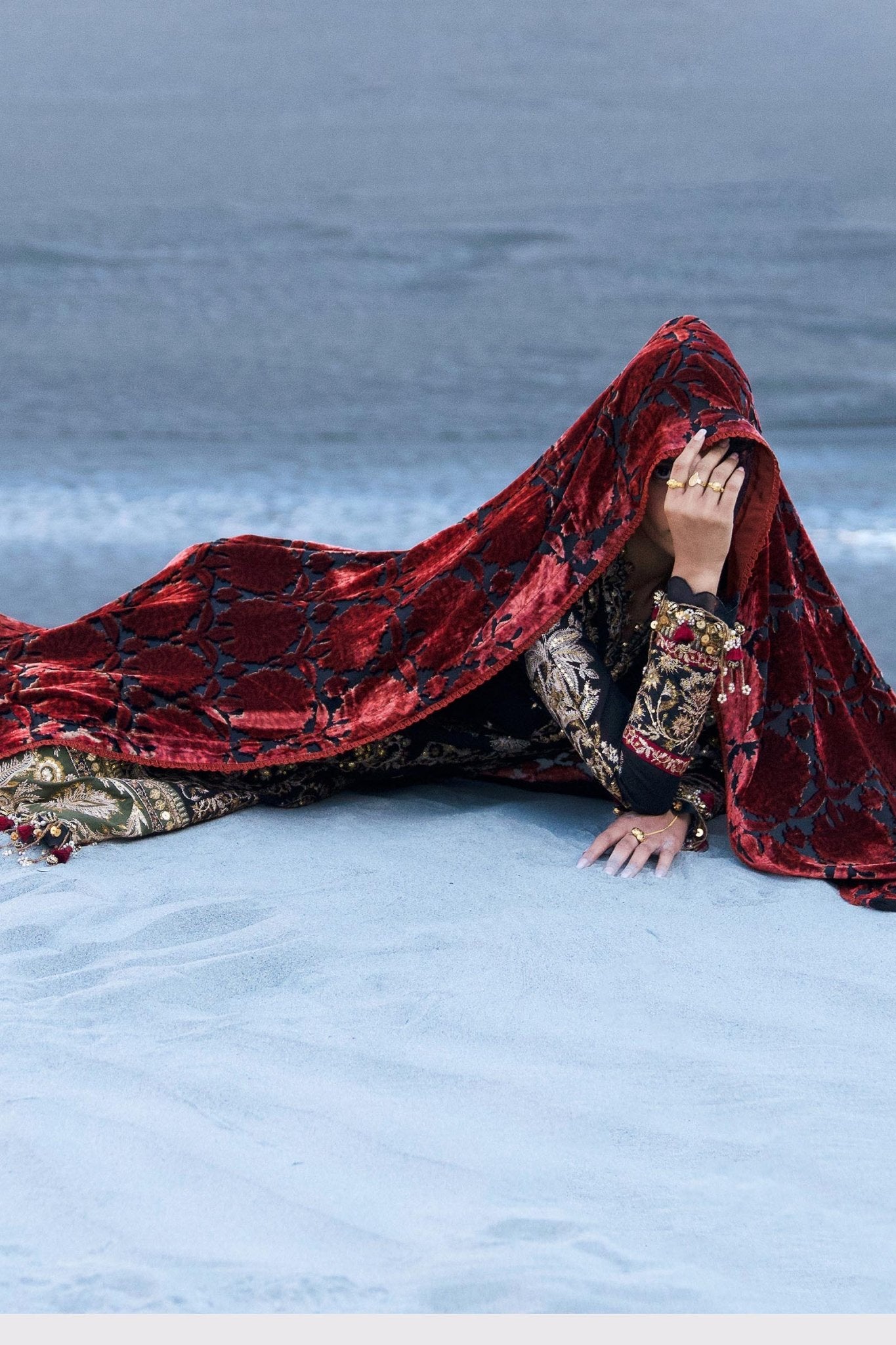 Model wearing Sana Safinaz Winter Luxury '24 S241 - 001A - 3CP, a black and gold embroidered outfit with intricate patterns and a bold red velvet shawl. Perfect for winter, available for Pakistani clothes online in the UK.